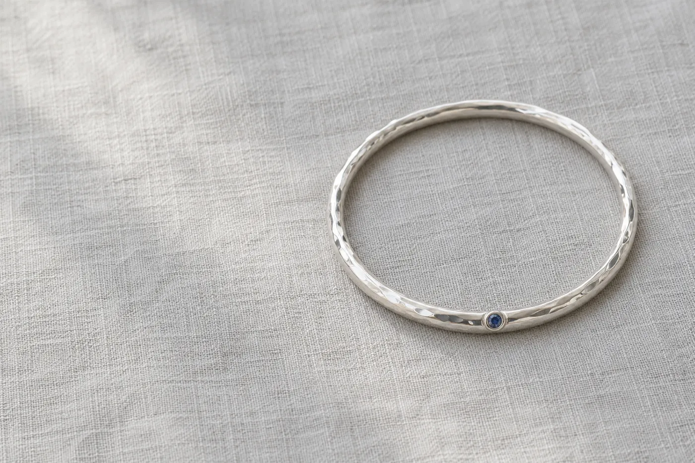 Illustration: gehämmerter Armreif aus Silber mit einem kleinen blauen Stein auf grauem Leinen